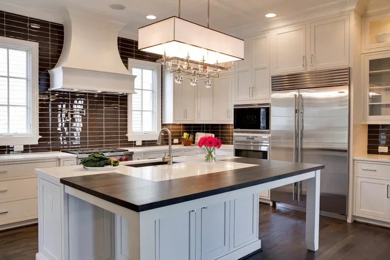 Dark glossy backsplash tiles surround a shaped white hood beside white cabinets and a rectangular shaded island light.