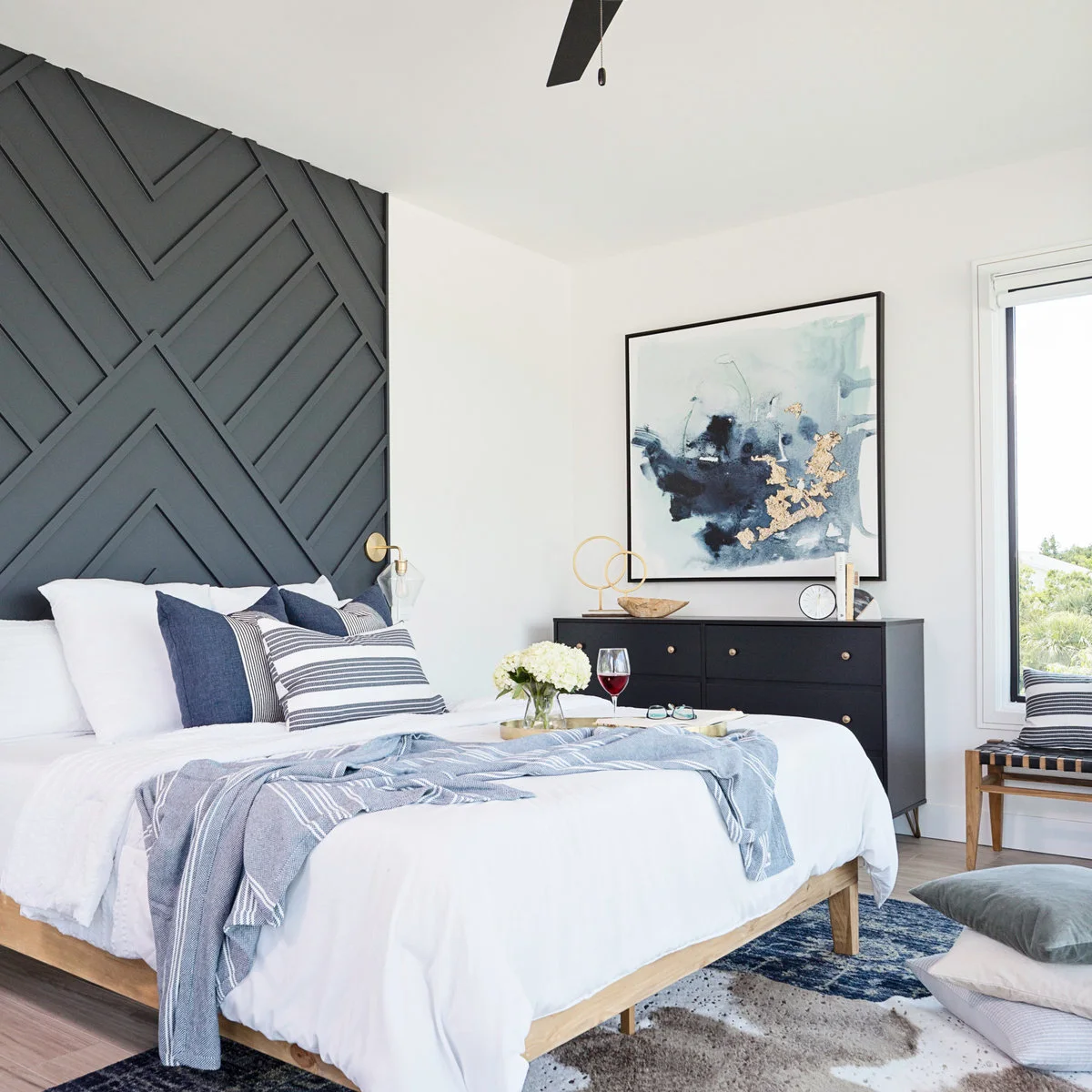 A navy, gray and cream abstract rug lies beneath a wood bed with blue pillows, beside a dark dresser and floor cushions.