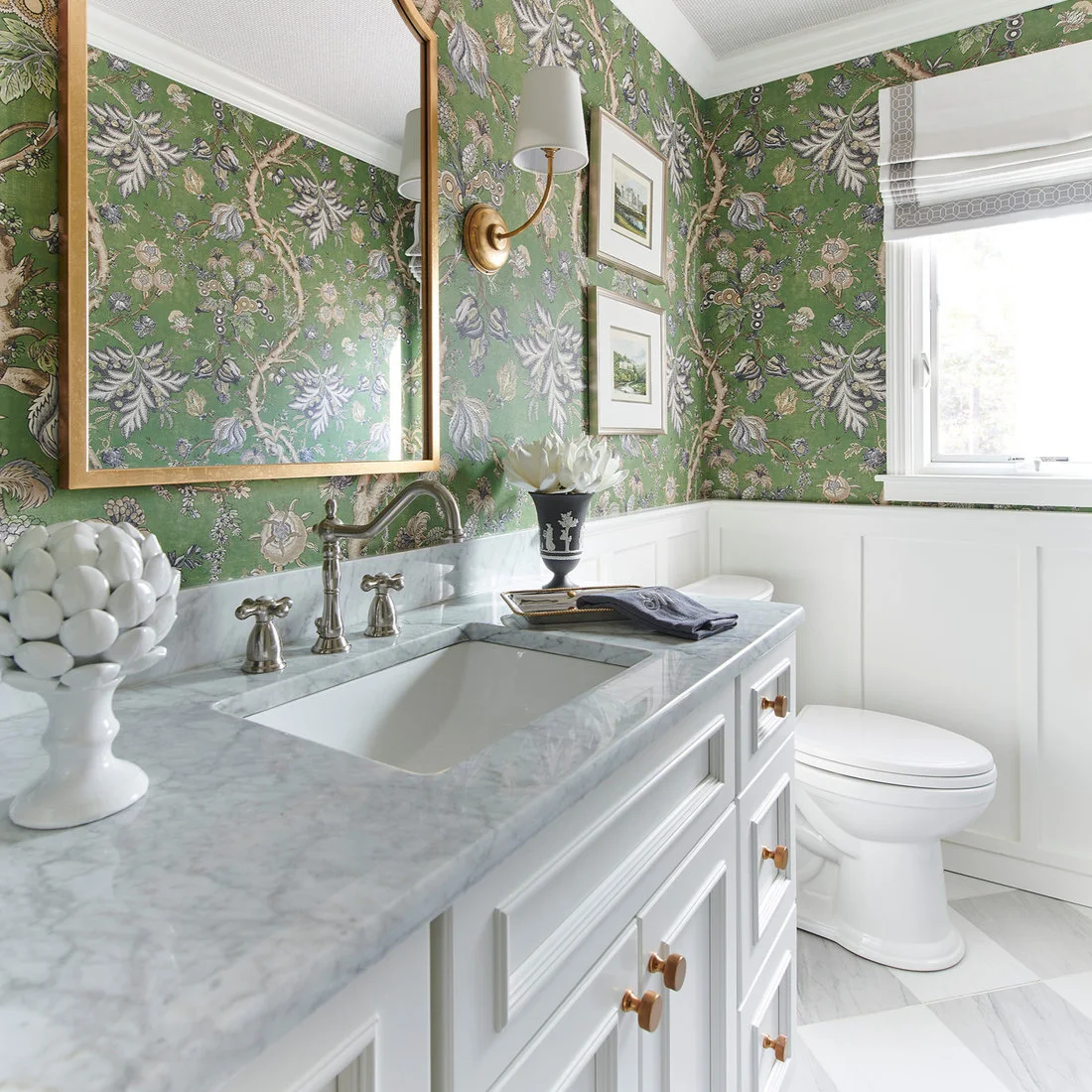 Green botanical powder-room wallpaper surrounds a gold-colored mirror and framed pictures above white paneling and a pale veined vanity counter