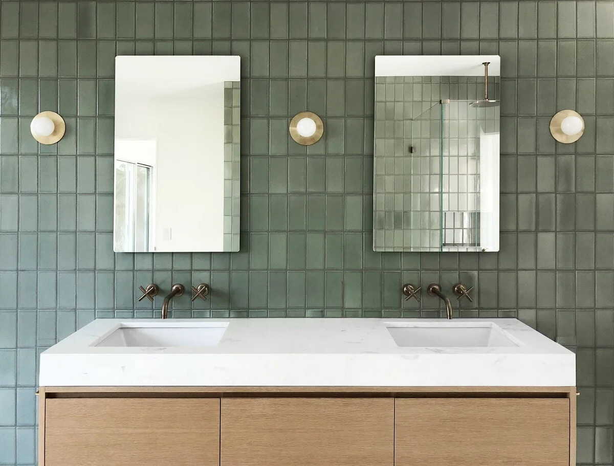 Sage rectangular tile surrounds two rectangular mirrors and a double white basin above a wood-front vanity with small round lights.