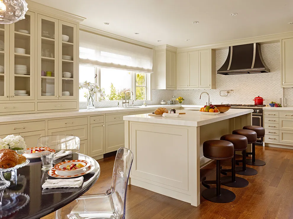 Beige cabinets with glass upper fronts surround a long island beneath a dark hood