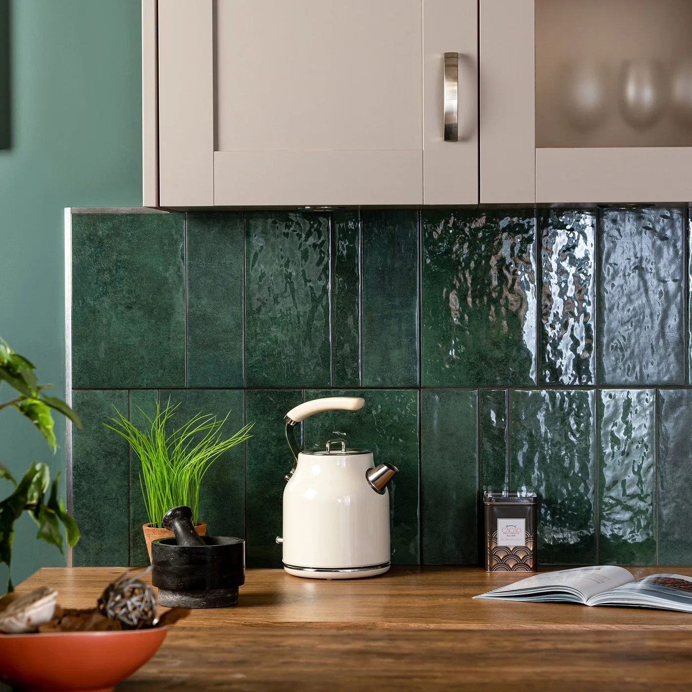 Deep green rectangular backsplash tiles sit above a wooden kitchen counter beside a cream kettle and pale wall cabinets