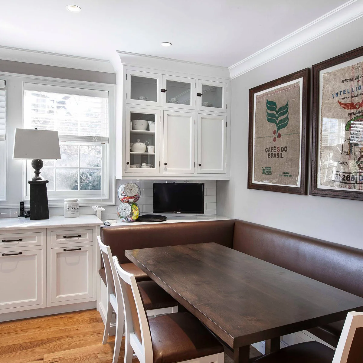 Two framed printed coffee sacks above a brown banquette beside white kitchen cabinets and a window.