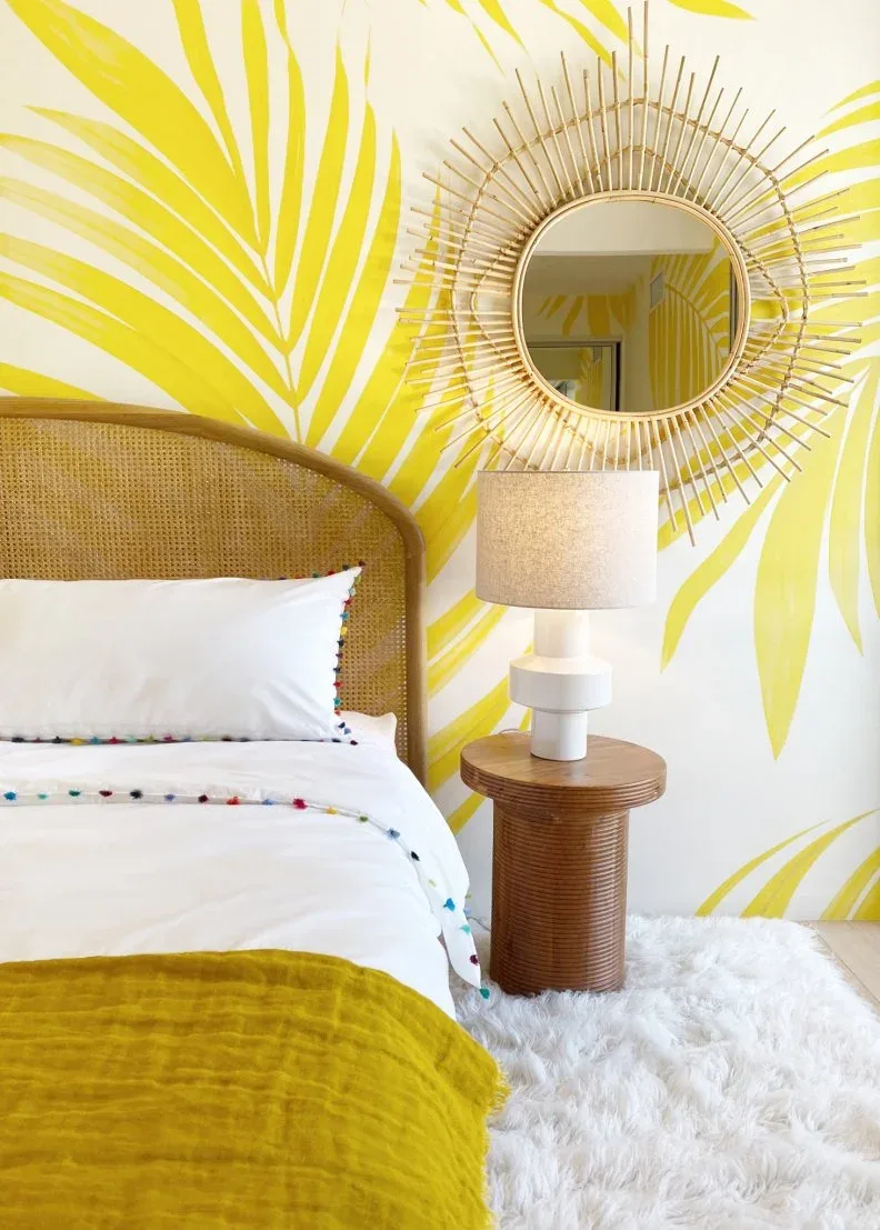 Bedroom detail with yellow leaf wallpaper, woven headboard, white bedding, yellow throw, wooden round table and sunburst mirror.