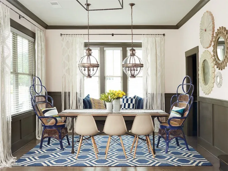 Pale openwork curtains with long fringe around dining-room windows with blinds, beside dark wall paneling, blue end chairs, white side chairs, and a geometric blue rug.