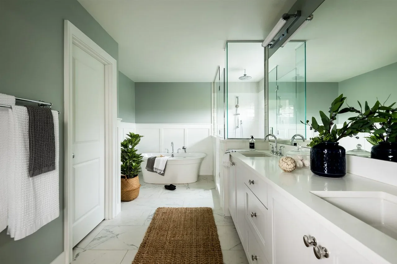 Soft gray-green upper walls sit above white paneling in a bathroom with a long white vanity, glass shower and freestanding tub.