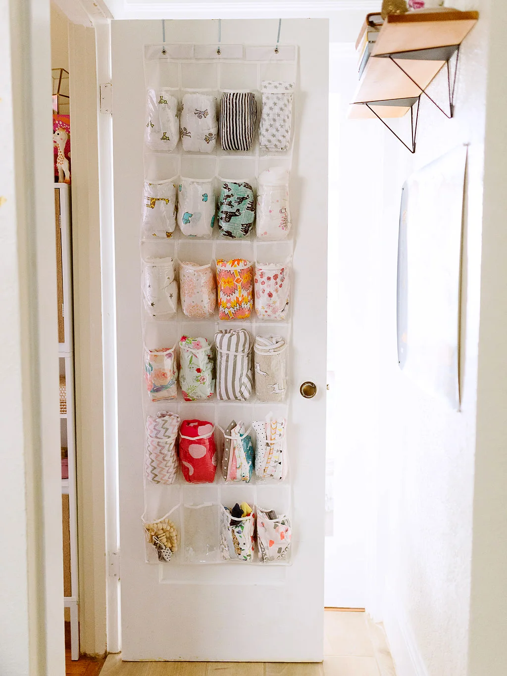 A clear pocket organizer hangs on the back of a white nursery door and holds rolled patterned textiles.