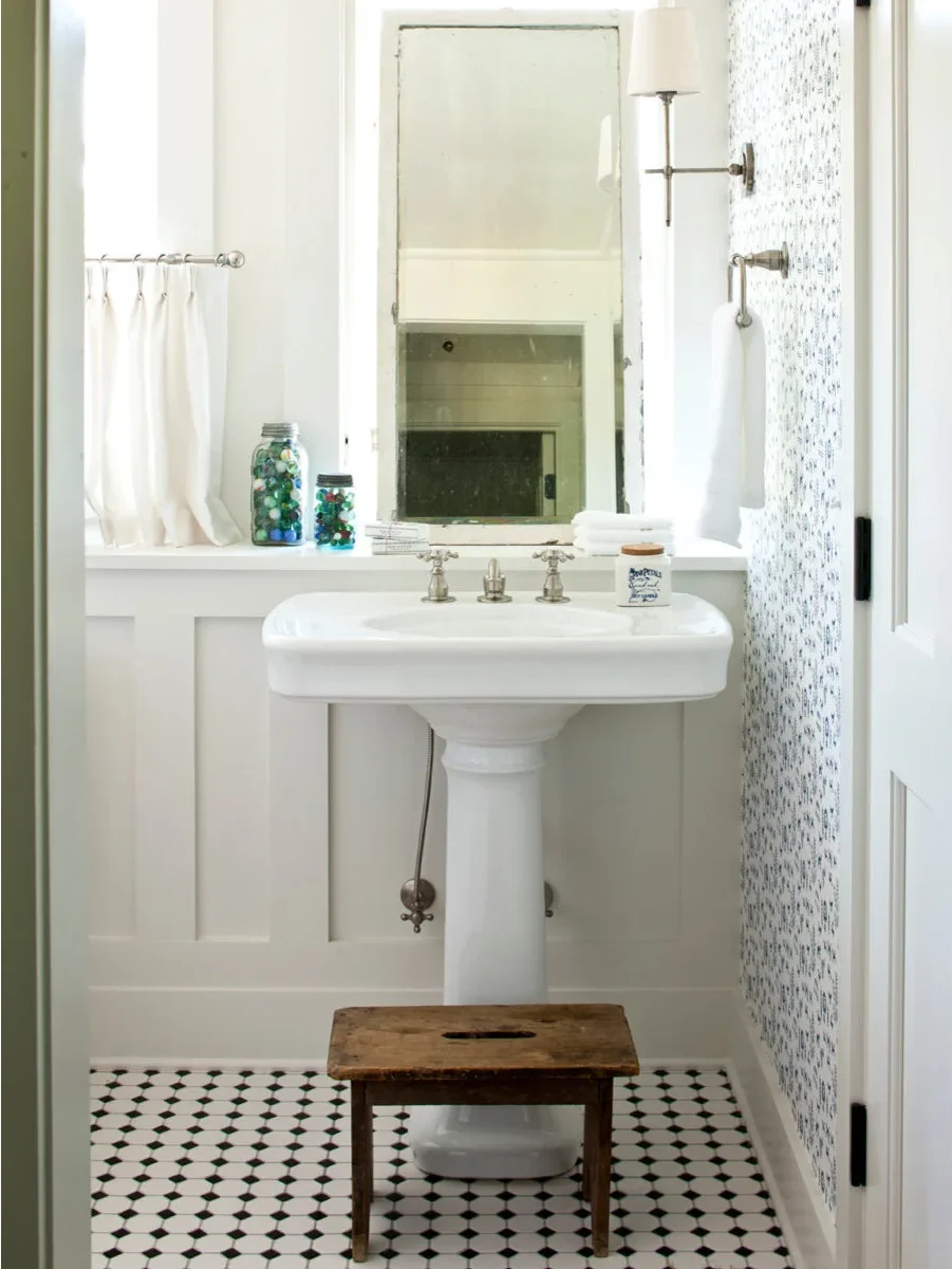 A white pedestal sink stands below a tall mirror with a shallow wall ledge behind the faucet and a small wood stool in front.