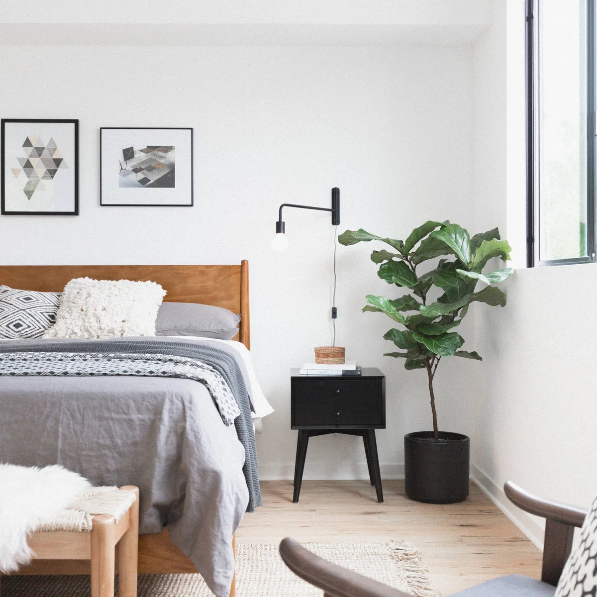 A bare globe lamp on a dark right-angle wall arm sits above a dark bedside table beside a wood headboard and tall leafy plant.