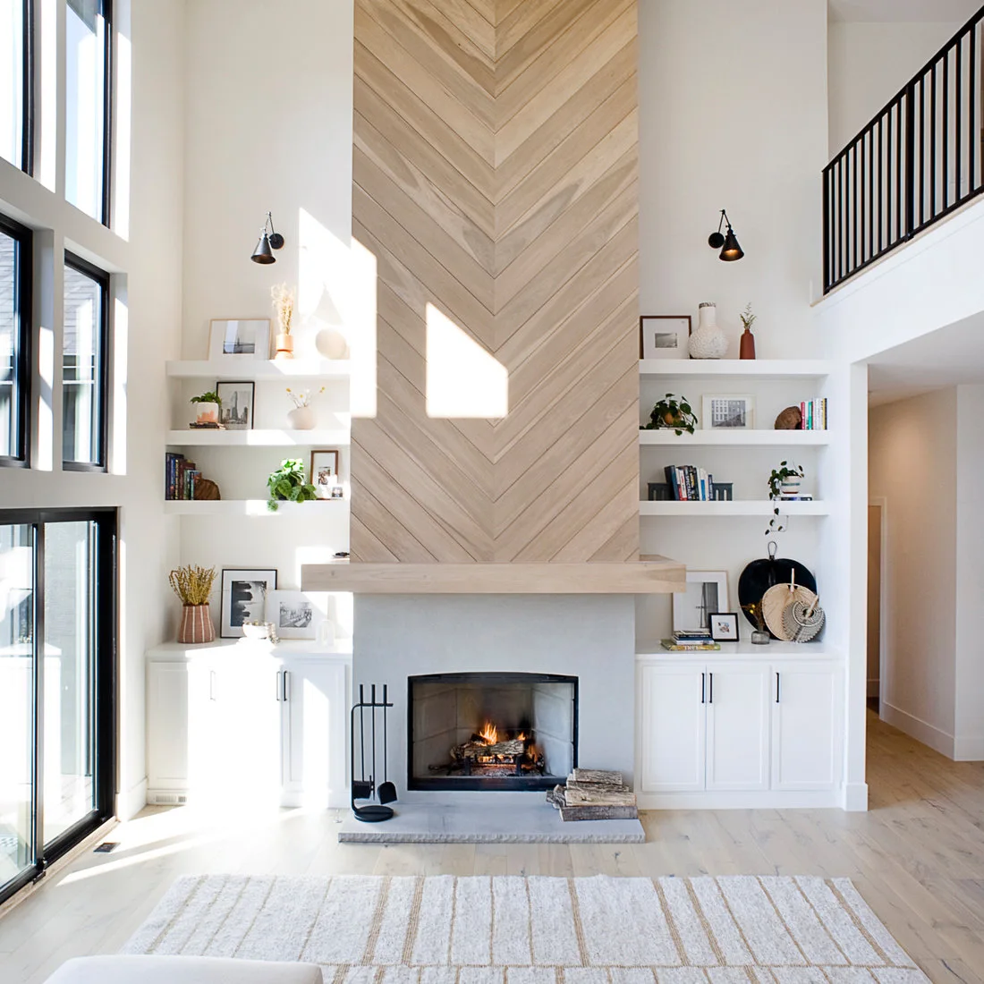 A tall pale chevron-patterned fireplace wall rises above a simple mantel and dark opening, with white shelves and lower cupboards on both sides and pale wood flooring.