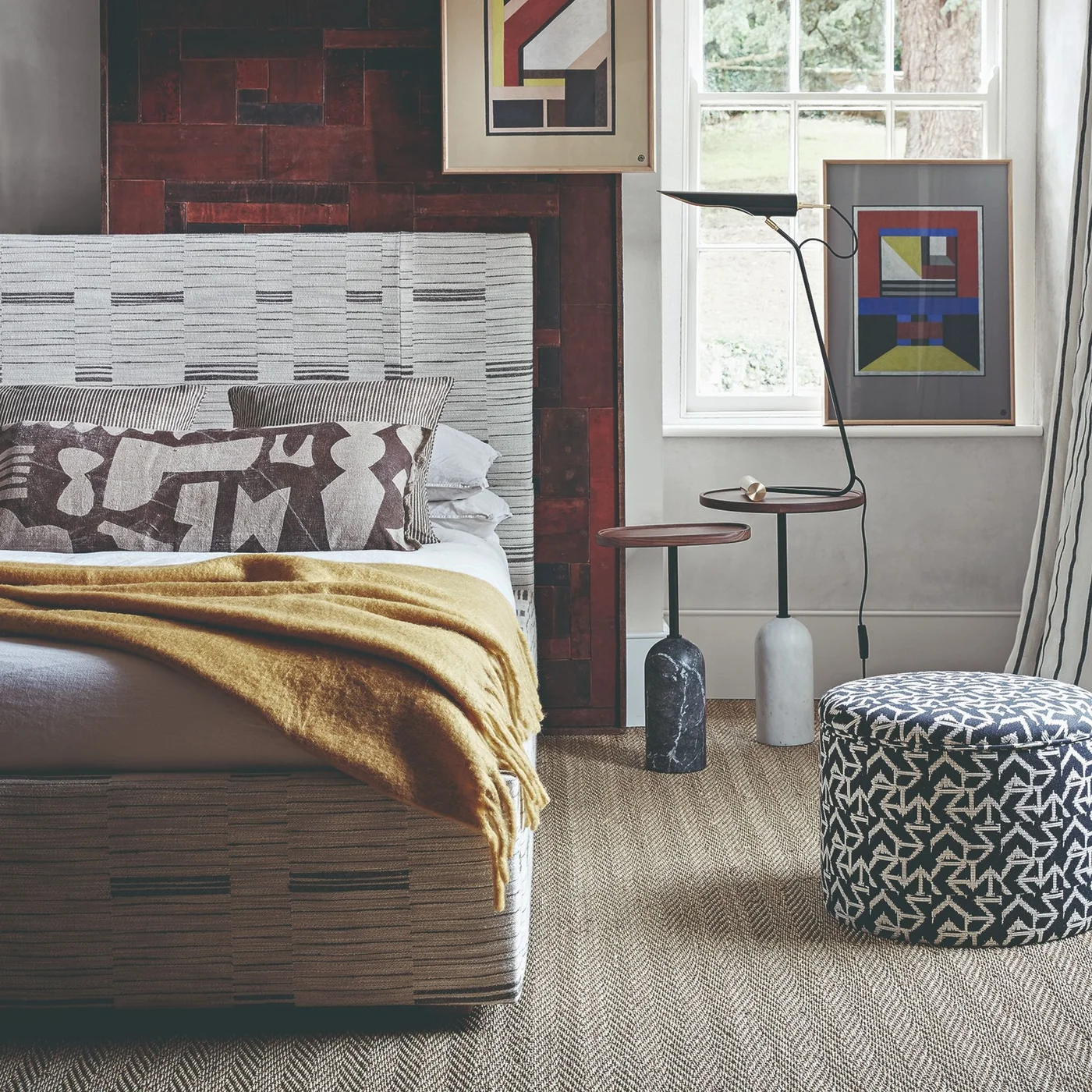 A patterned upholstered bed sits beside a narrow two-level pedestal stand and an angled reading lamp, with geometric art and a patterned stool nearby.