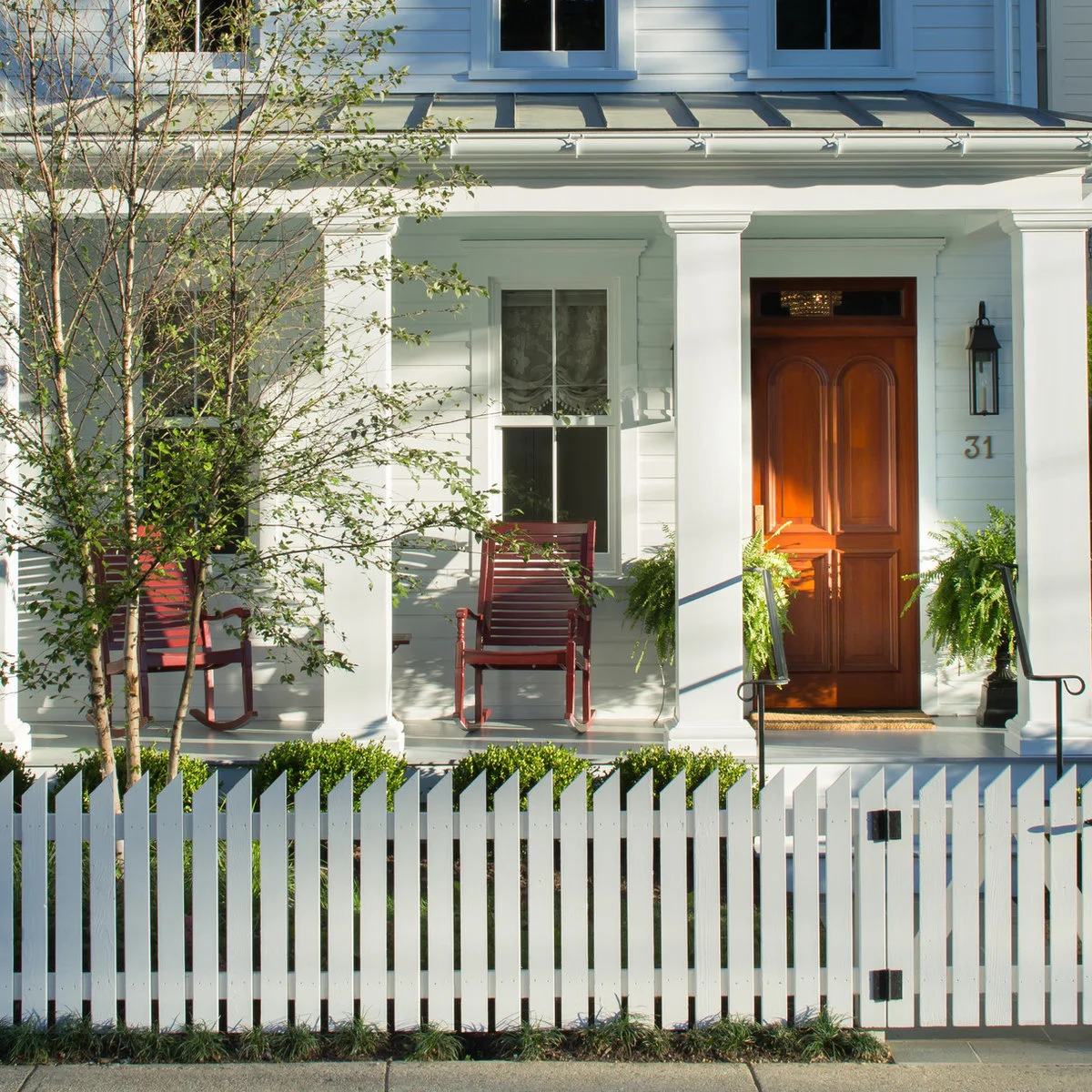 Shallow white front porch with red rocking chairs and greenery beside the wood front door