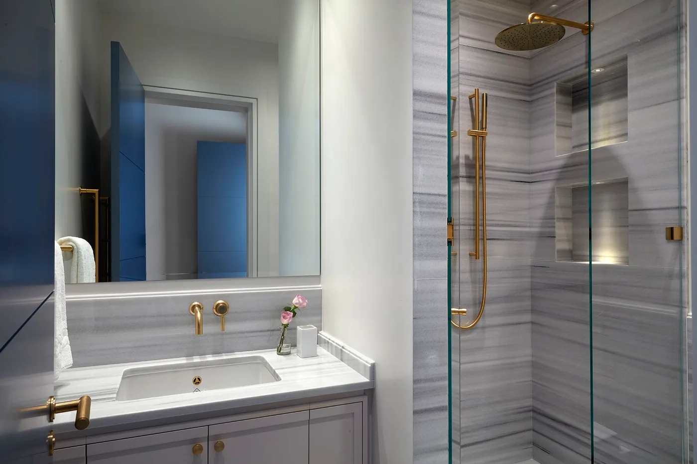 A navy bathroom door and its reflection introduce blue beside gray veined tiles, a pale vanity and a glass shower