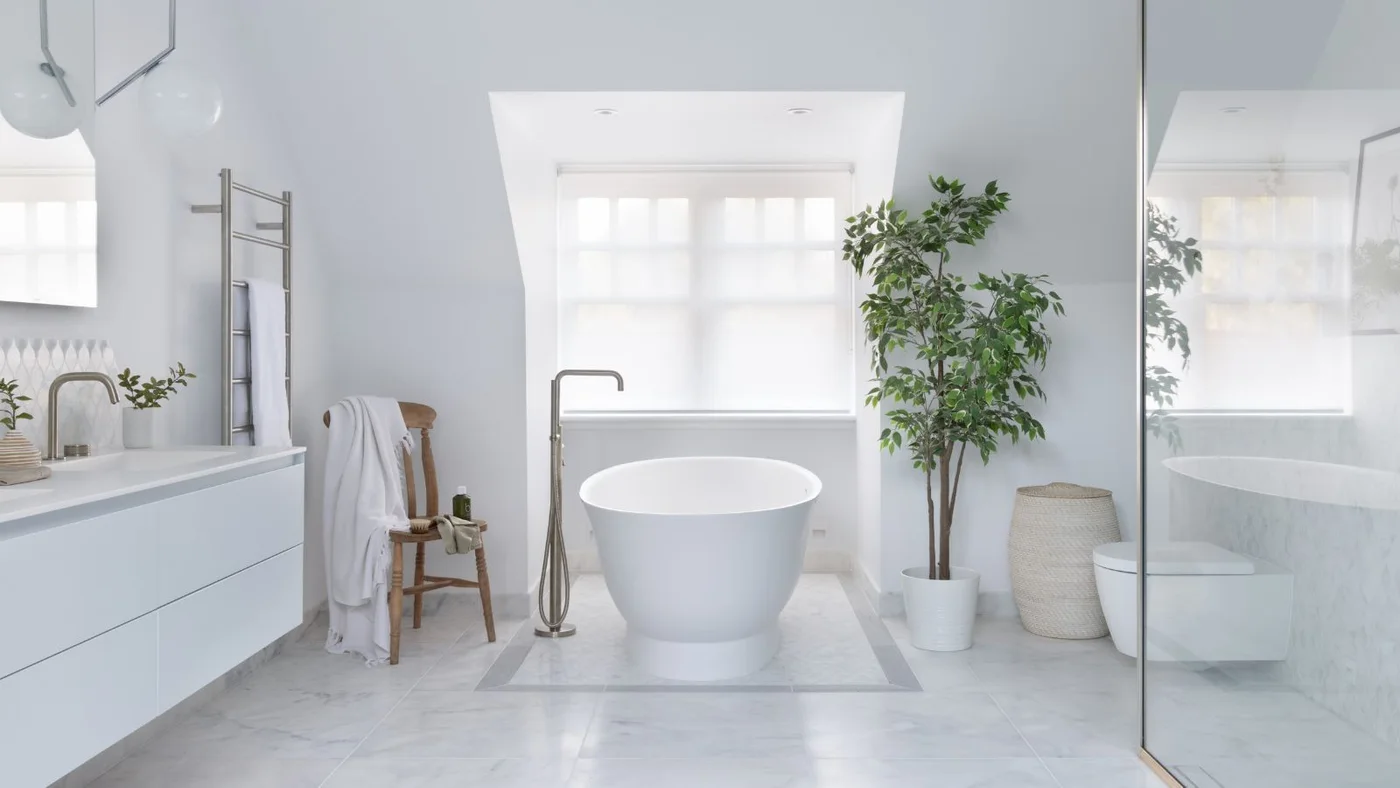 A white oval bathtub stands beneath a pale window beside a flat-fronted white vanity, glass shower screen, wood chair and leafy plant