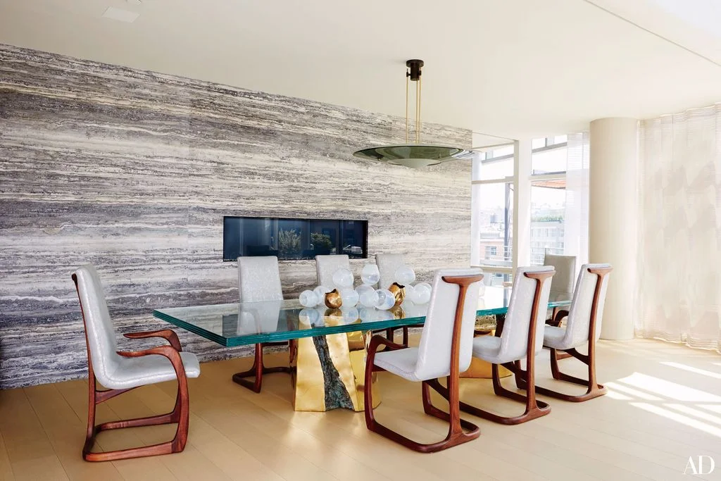 A narrow dark fireplace opening sits in a broad gray horizontally patterned wall behind a glass dining table, pale upholstered chairs and a round hanging light