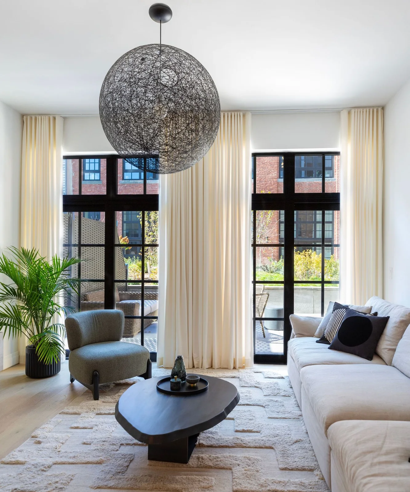 A dark irregular coffee table sits on a textured pale rug between a white sofa, gray chair and tall cream curtains.