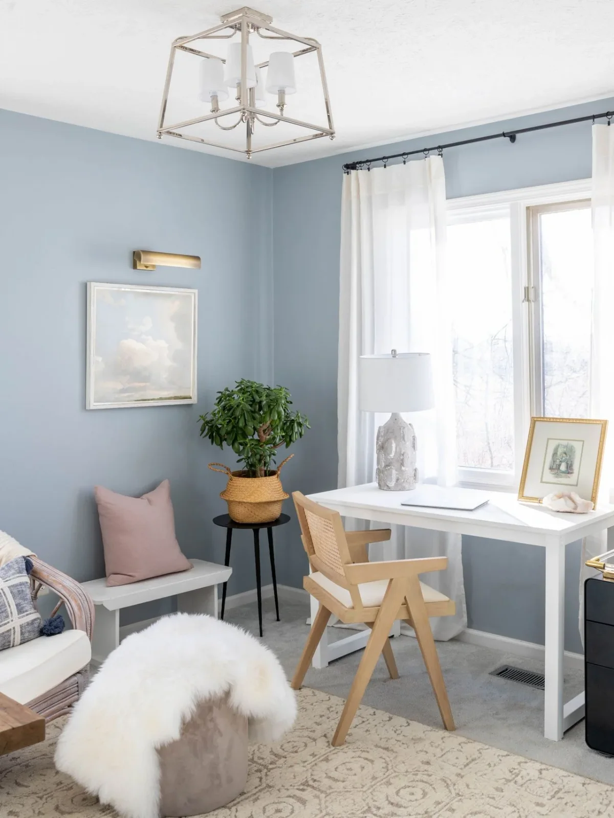 A pale blue office corner contains a white desk, wood chair, white curtains, a narrow bench and a small dark drawer unit.