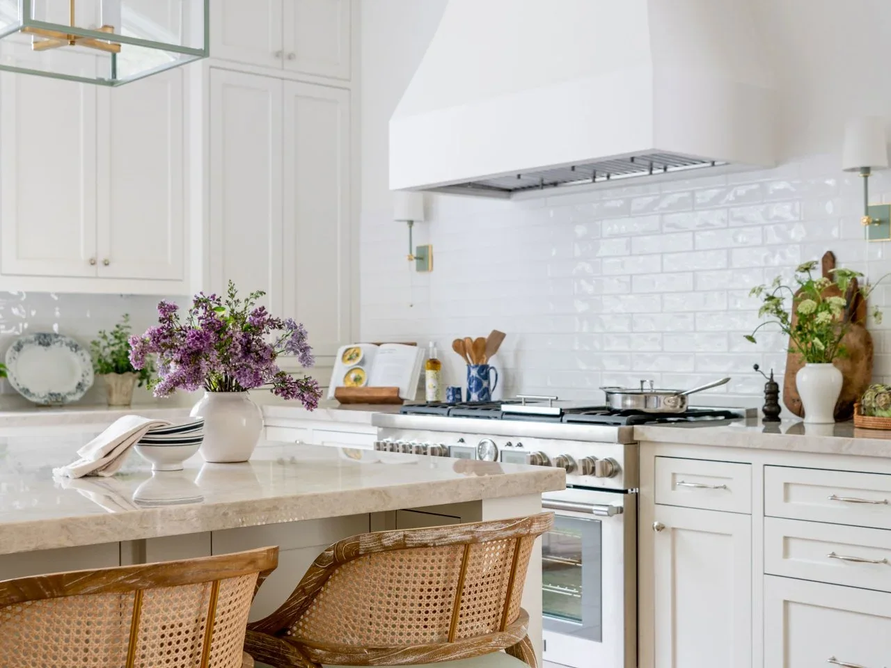 Glossy white subway tiles fill the wall beneath a broad white kitchen hood beside pale cupboards and woven stools.