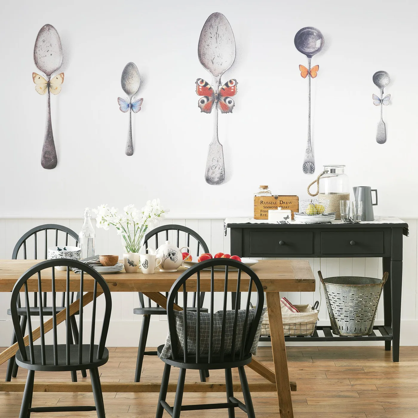 Country dining room with black spindle chairs, wooden table and bench, dark side table and large spoon-and-butterfly wall decoration.