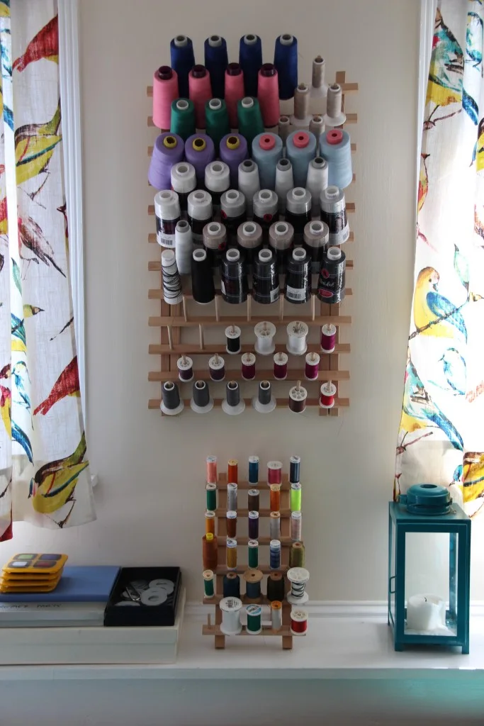 Rows of large thread cones sit on a wall-mounted rack between patterned curtains, with smaller spools on a lower stand.