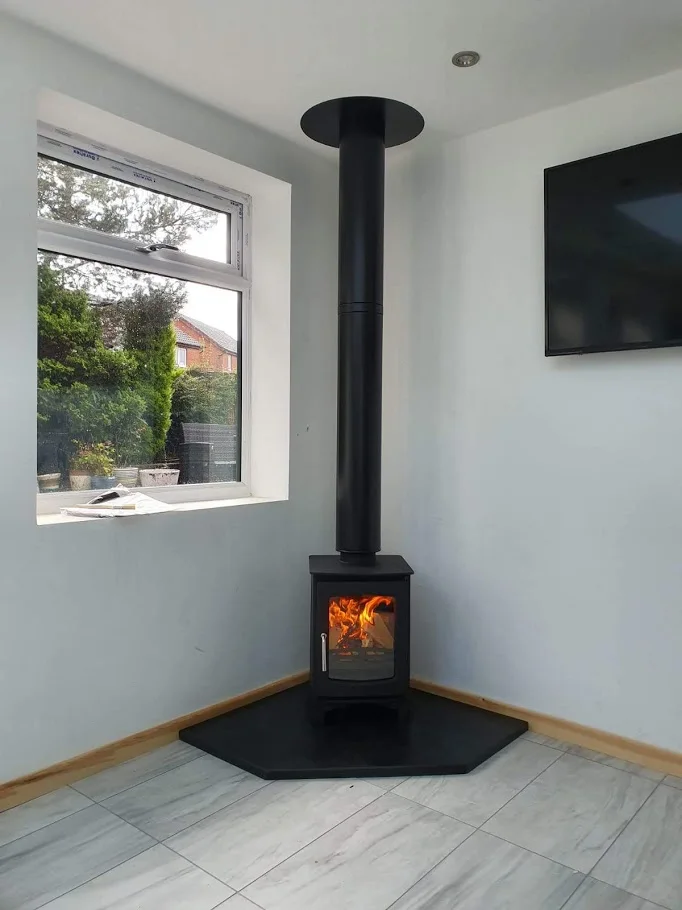 A black corner stove and vertical flue stand on a five-sided slate hearth beside pale plank-pattern flooring.
