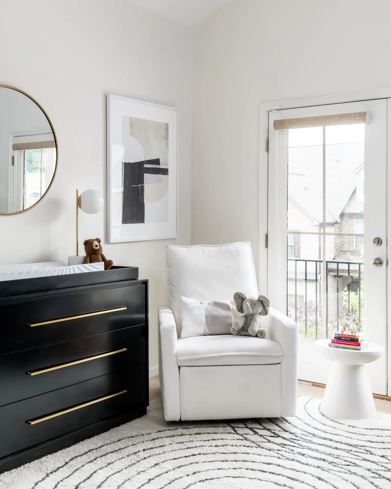 A pale nursery adult chair sits beside a dark dresser with warm-colored handles below a framed graphic print near glazed doors and a small white table