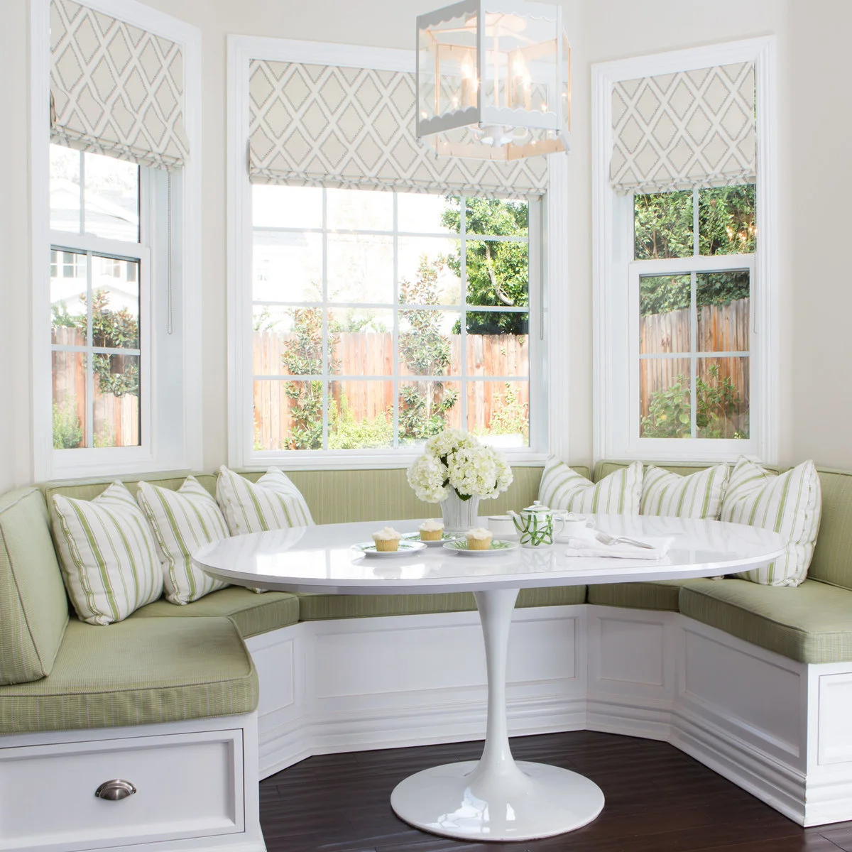 A green upholstered bay banquette with striped pillows, white pedestal table and geometric Roman blinds.