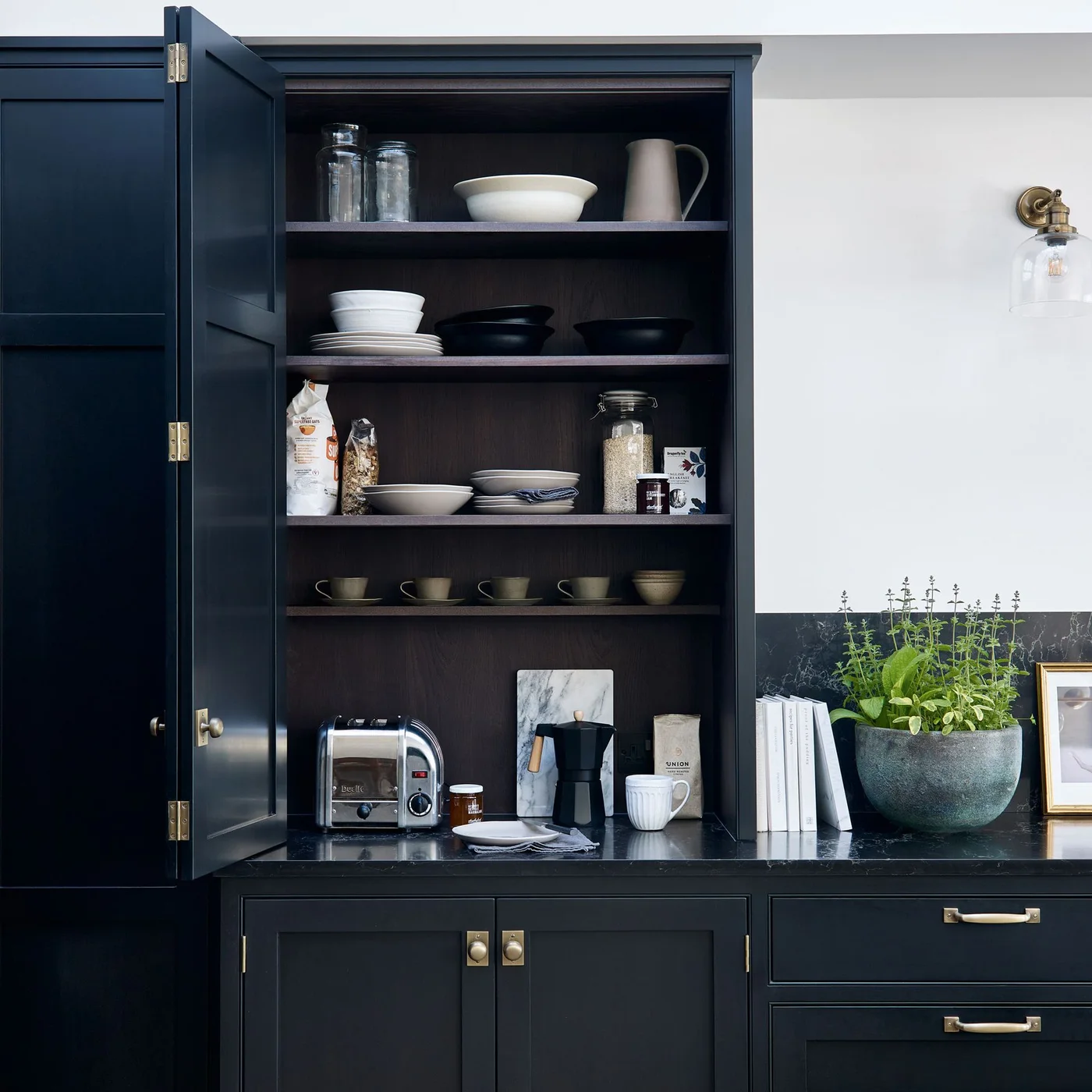 An open dark kitchen cabinet holds dishes, jars, mugs and small appliances on shelves beside a dark counter, pale wall and warm-colored hardware