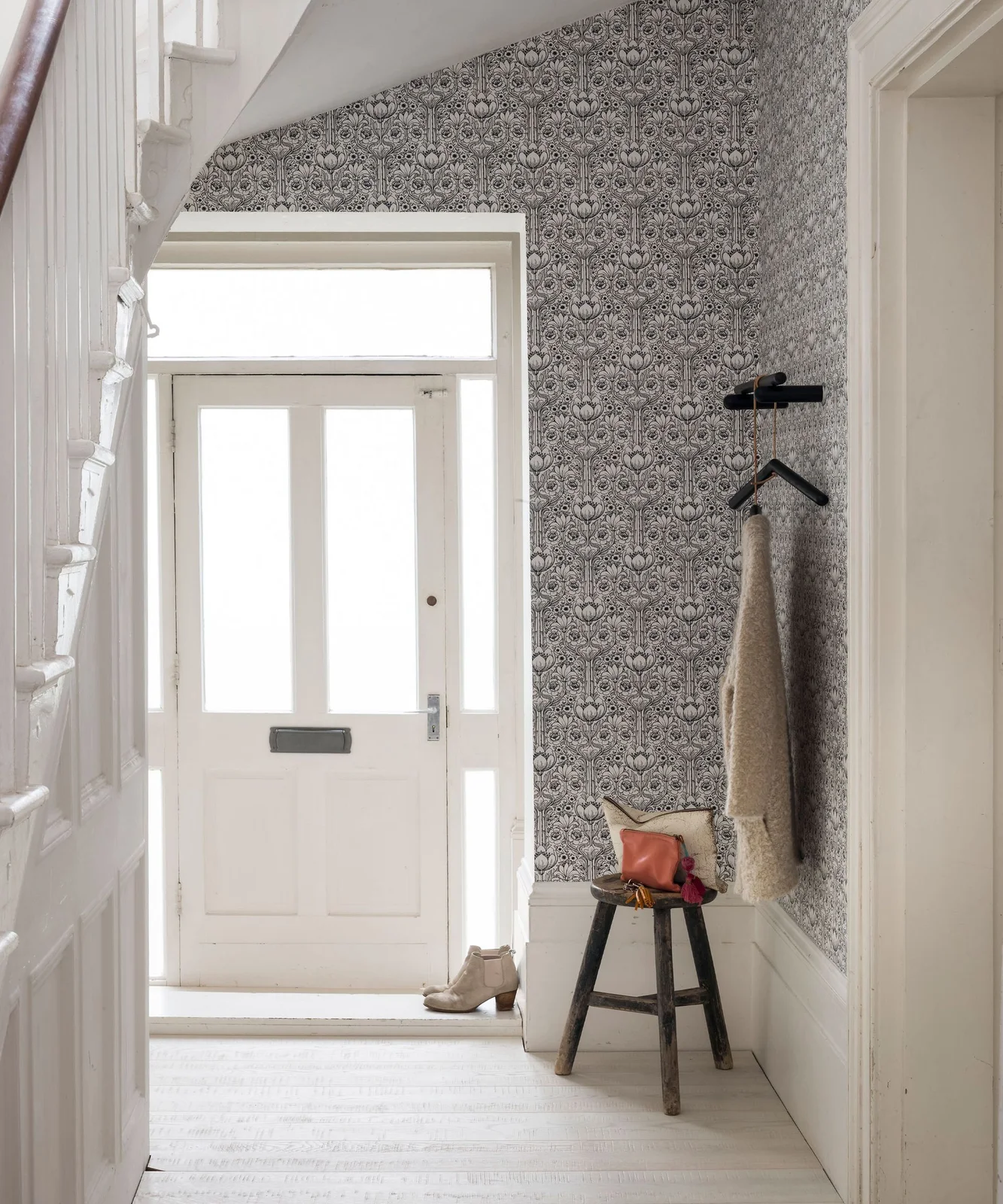 A white front door stands beside fine dark botanical wallpaper, a small dark wall rail, a hanging coat and a wooden stool above pale flooring