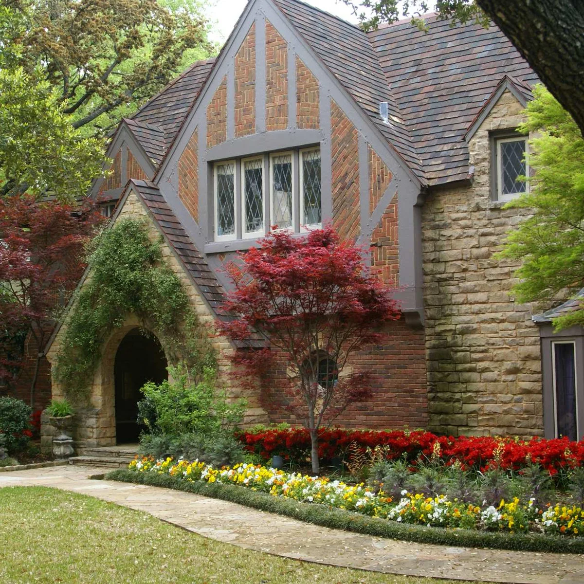 A low dark-green planted edge follows a stone path in front of yellow and white flowers and a stone-and-brick house.