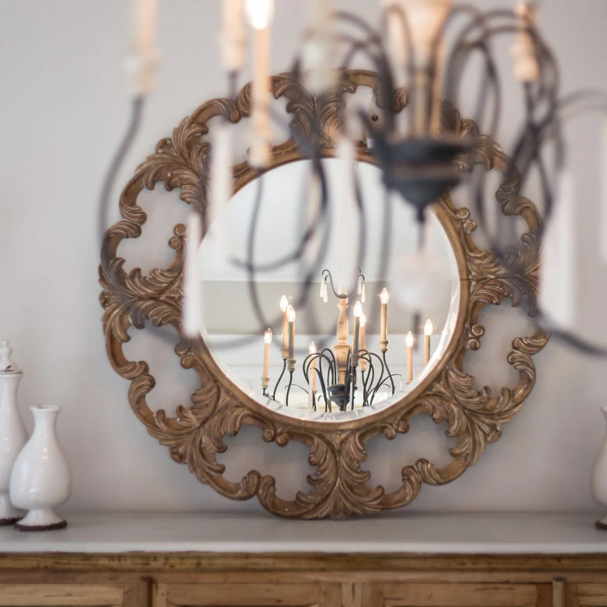 Ornate round gold-colored mirror above a pale cabinet reflecting a chandelier