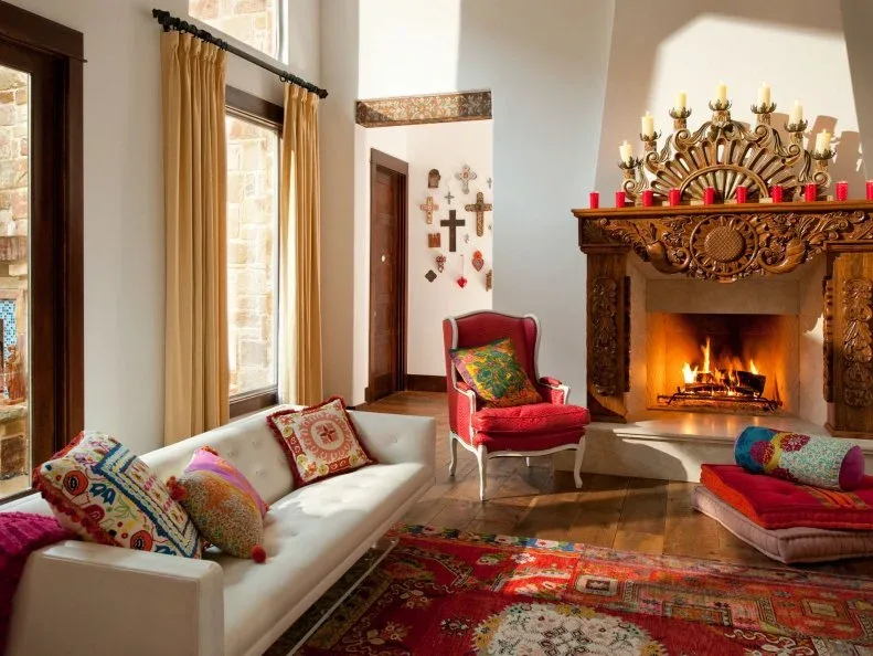 Carved dark wood fireplace mantel in pale room with tall curtains, colorful cushions and patterned rug