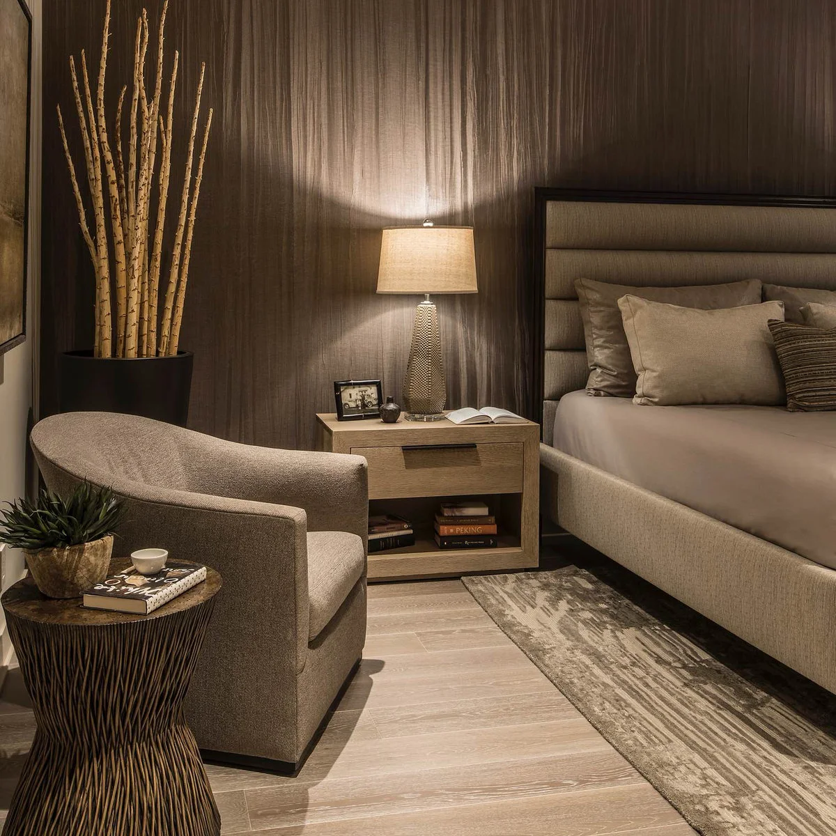 Textured brown wall behind a neutral upholstered bed with a brown chair and a wooden bedside table.