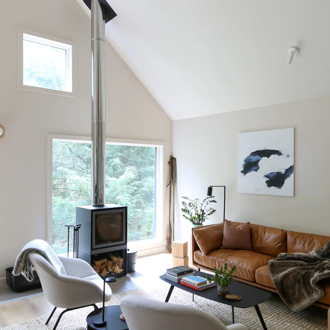 A black square stove with a tall metal flue stands before large windows beside a brown sofa, two pale chairs and a low dark coffee table.