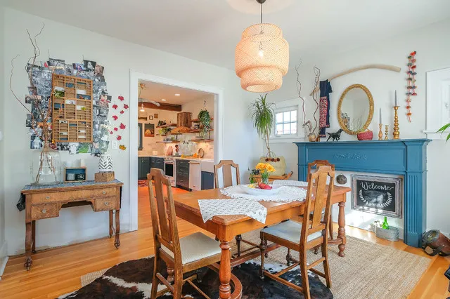 A timber table and mismatched chairs sit beside a blue-painted fireplace, wall art and a round mirror.