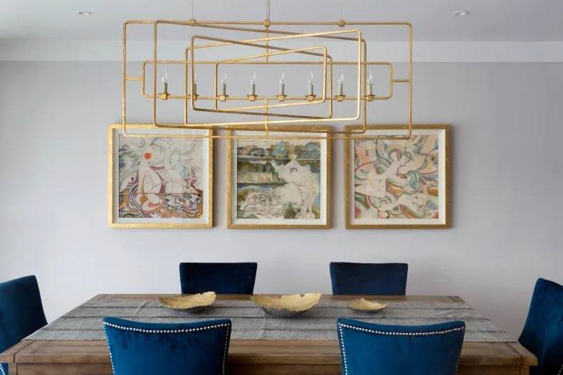 Three low gold-toned bowls across a wood dining table with deep blue chairs beneath a geometric chandelier