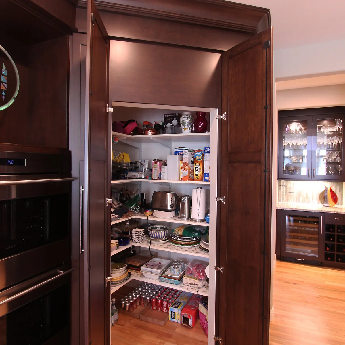 Dark paneled double doors open beside ovens to reveal white pantry shelving holding dishes, packets and small appliances.