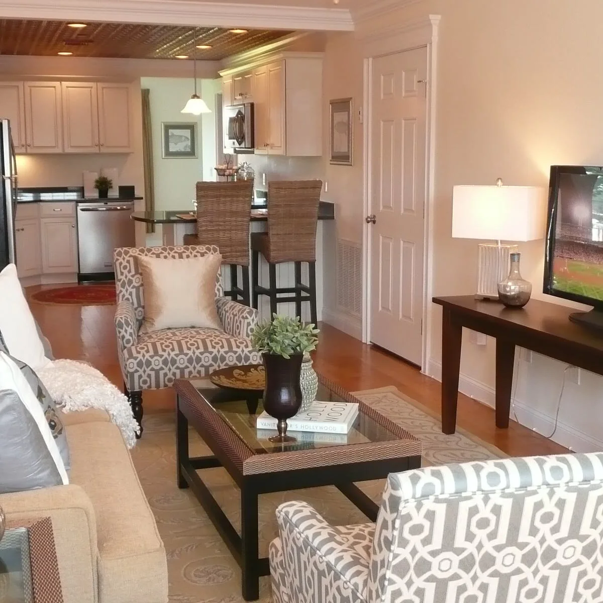 Long living area with a pale sofa, patterned chairs, glass coffee table and kitchen breakfast stools behind