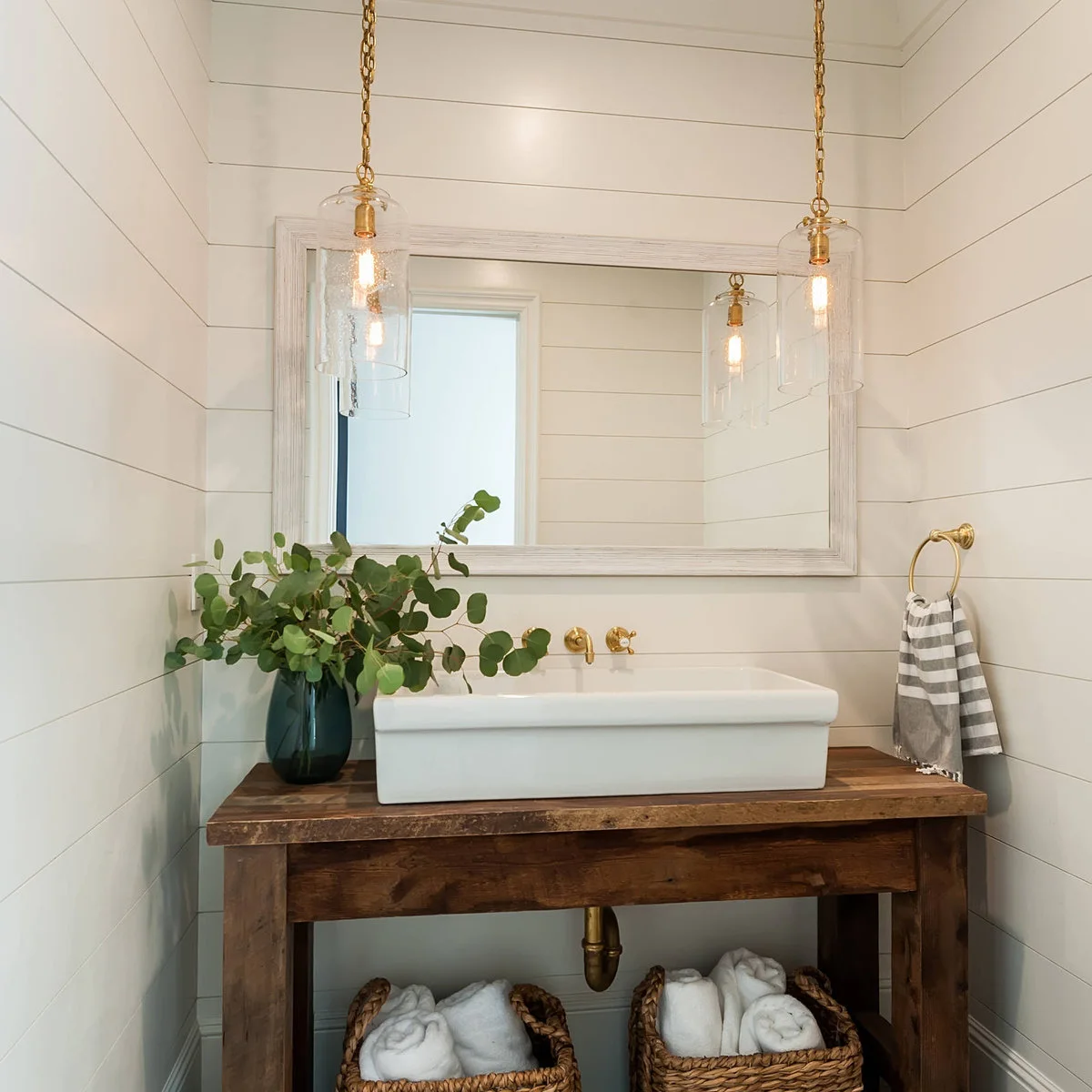 A white rectangular vessel basin on a rustic wood console with baskets below and glass pendants above.