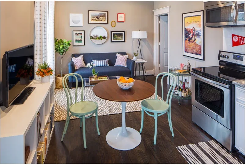 A small round wood-topped pedestal table and two pale green chairs sit between a kitchen run, a low television unit and a blue sofa.