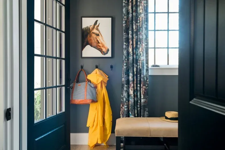 Dark blue-gray entry with a deep teal glazed door, floral curtain, horse portrait, wall hooks, yellow raincoat, and tan upholstered bench.