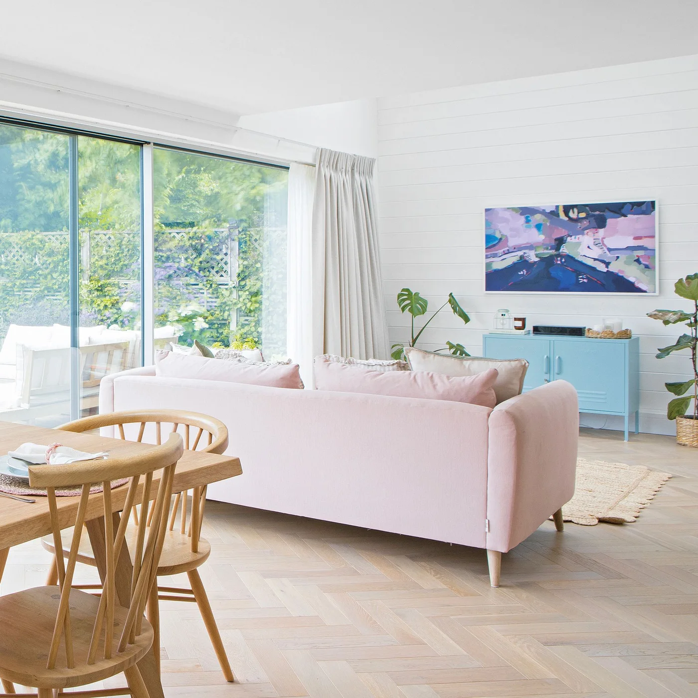 A pale pink sofa separates dining chairs from a sitting area with a blue cabinet and large garden glazing.