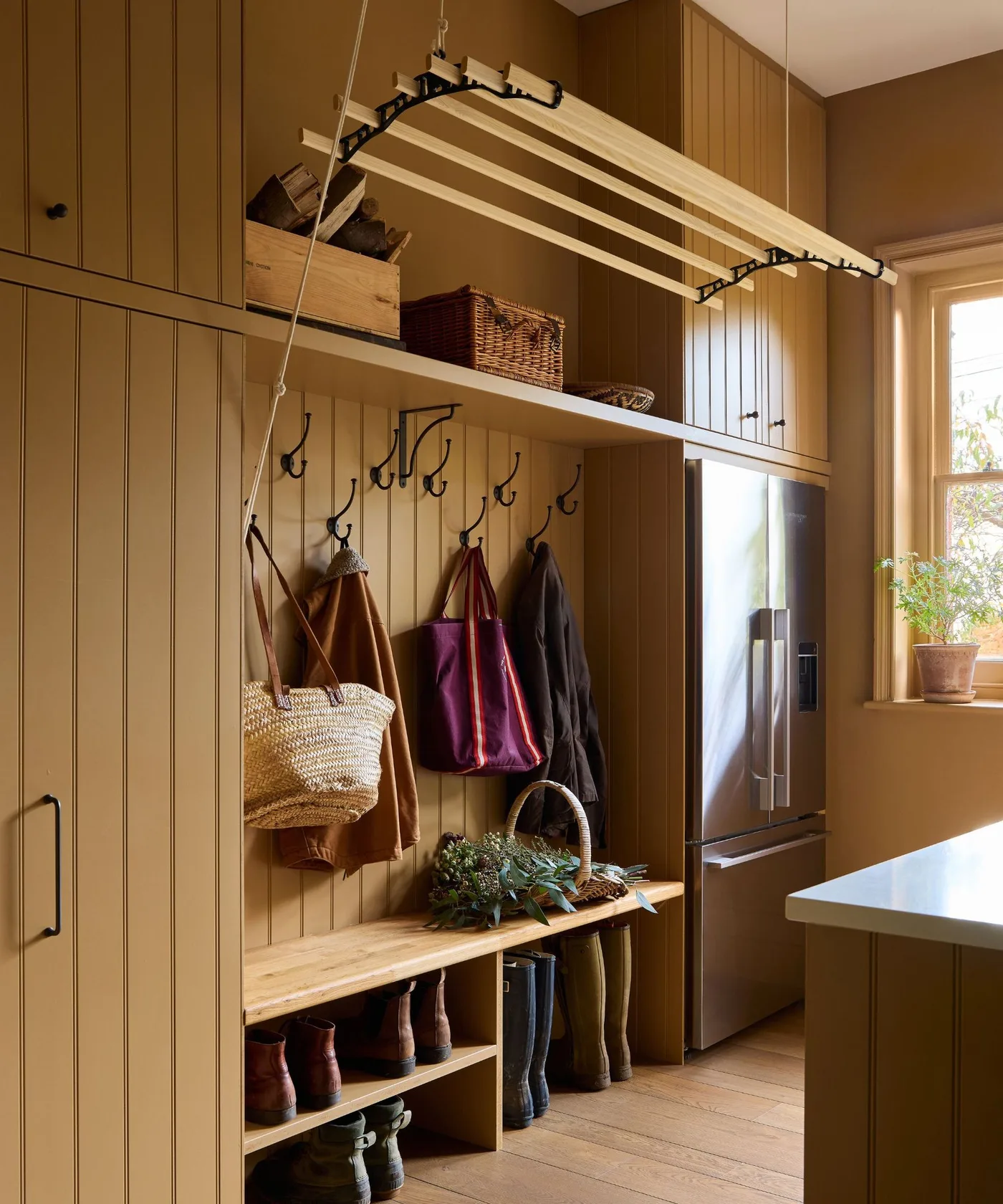 Brown paneled cabinetry surrounds a wood boot bench, coat hooks and open shoe shelves beneath a suspended slatted drying rack beside a refrigerator.