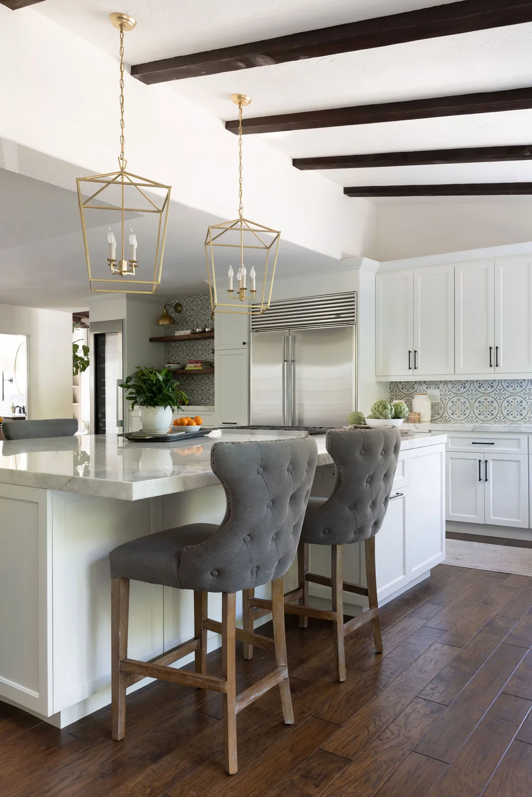 A white kitchen has blue-gray patterned backsplash tiles, dark ceiling beams, warm metal lanterns and gray upholstered stools at a paneled island.