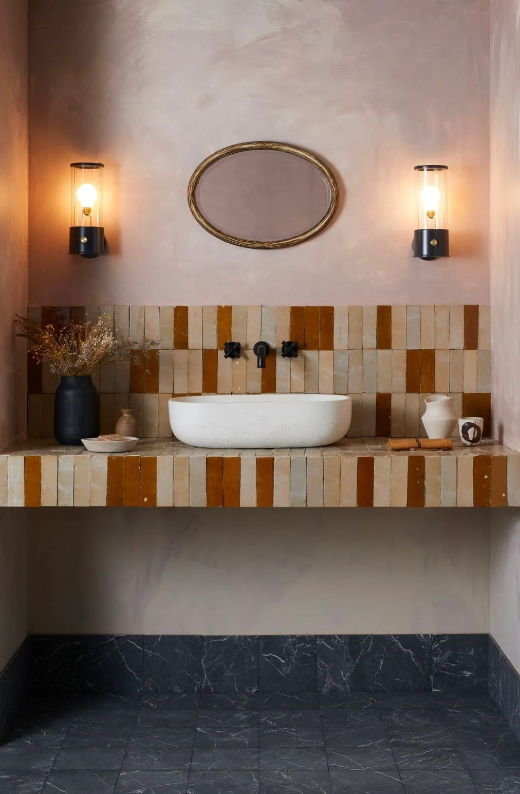 A white oval vessel basin stands on a warm striped tiled shelf below a small oval mirror between two cylindrical wall lights against a pale pink wall