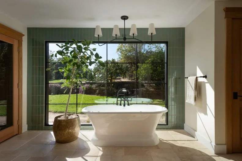 A white freestanding tub sits beside a green tiled wall and broad dark-framed window beneath a shaded hanging light with a large potted plant nearby