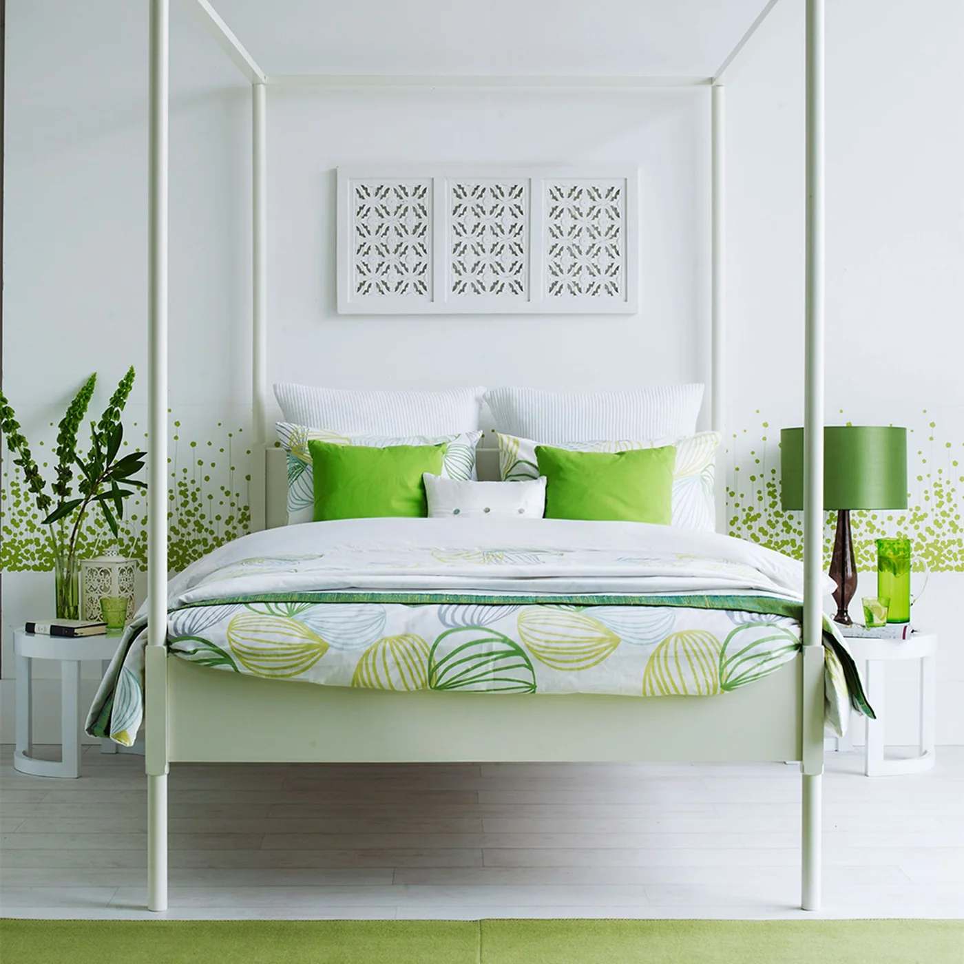A pale four-poster bed has bright green cushions and patterned bedding beside a green lamp and small leafy accents on white walls