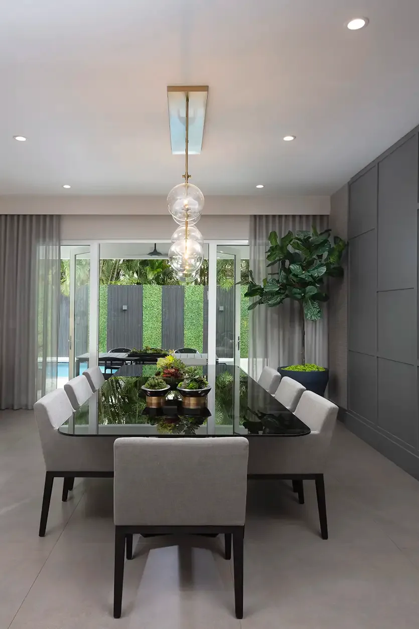 Dark glass dining table surrounded by pale upholstered chairs beneath clear globe pendants against a gray paneled wall