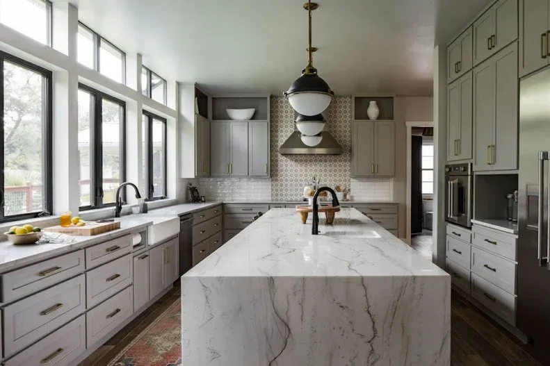 Gray-green kitchen cabinets around a pale veined island, black-framed windows, dark pendants and patterned tile behind the range