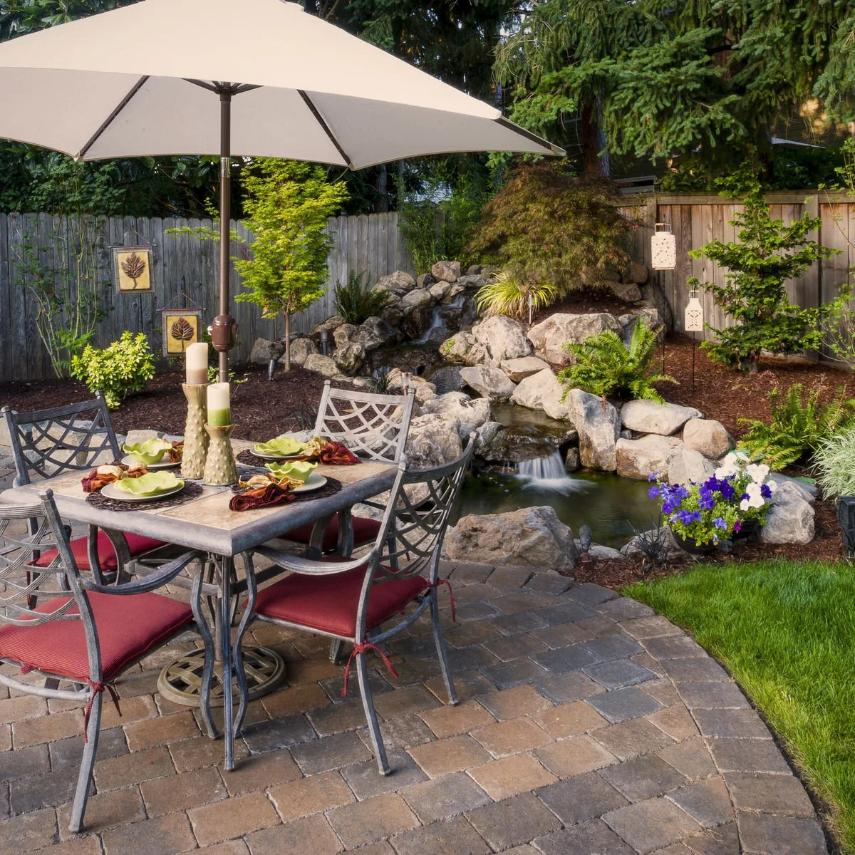 A garden dining table beside a compact rock waterfall and small pond, with a wood fence and mixed green planting behind.